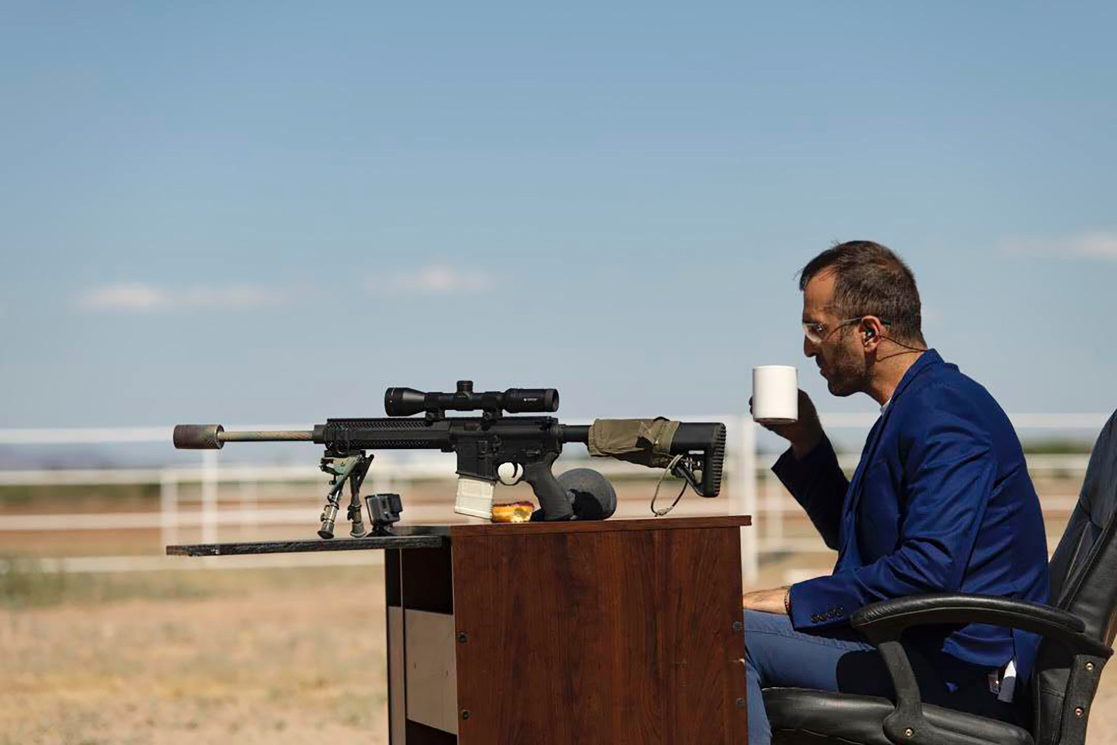 man with rifle drinks coffee at a desk in the desert
